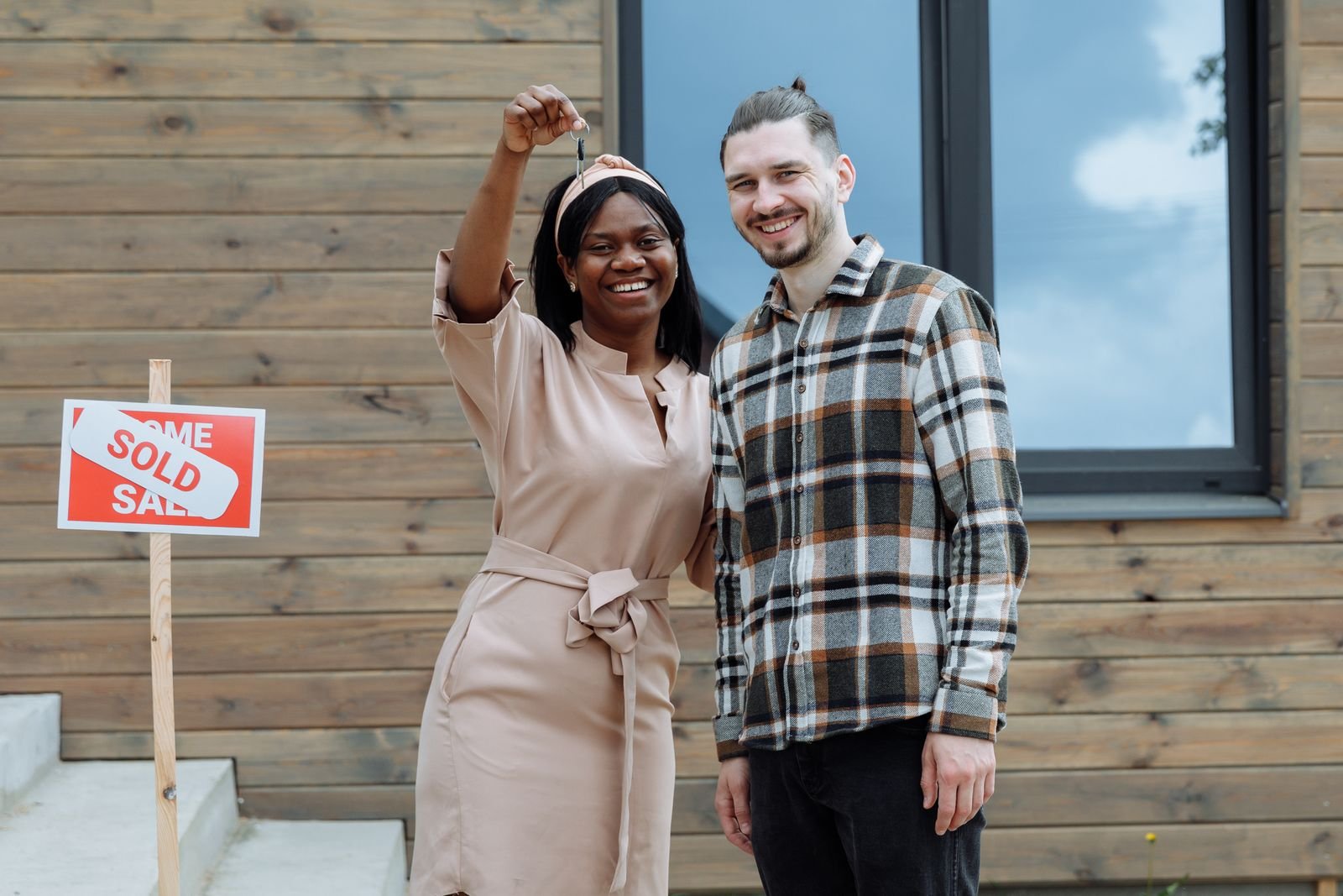 Happy first-time home buyers holding up their new house keys beside a sold sign